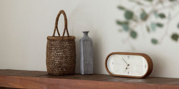 Woven basket, ceramic vase, and clock on a wooden surface with a blurred plant in the background.